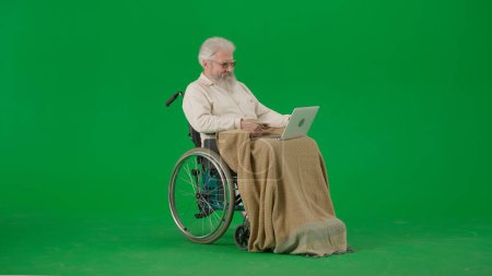 Photo for Pensioner everyday life creative advertisement concept. Portrait of aged man isolated on chroma key green screen background. Senior man in wheelchair covered by plaid holding laptop. - Royalty Free Image
