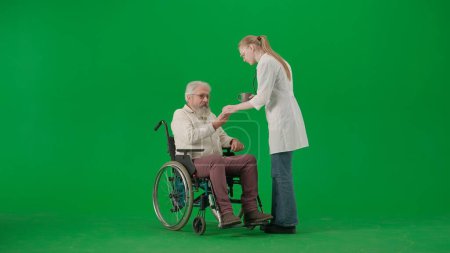 Photo for Pensioner everyday life creative advertisement concept. Portrait of aged man isolated on chroma key green screen background. Senior man in wheelchair taking pills and water from a nurse. - Royalty Free Image