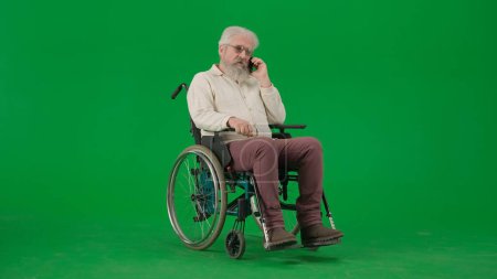Photo for Pensioner everyday life creative advertisement concept. Portrait of disabled man isolated on chroma key green screen background. Senior man sitting in wheelchair talking on smartphone, full shot. - Royalty Free Image