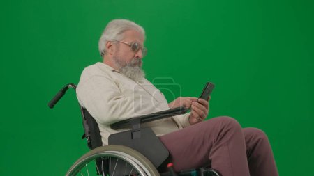 Téléchargez les photos : Pensionné vie quotidienne concept publicitaire créatif. Portrait d'un homme handicapé isolé sur écran vert chromatique de près. Homme âgé assis dans un fauteuil roulant tenant smartphone regarder la vidéo. - en image libre de droit