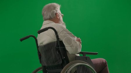 Photo for Pensioner everyday life creative advertisement concept. Portrait of disabled man isolated on chroma key green screen close up. Senior man sitting in wheelchair with his back at the camera, looking up. - Royalty Free Image