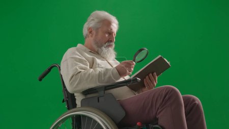 Téléchargez les photos : Pensionné vie quotidienne concept publicitaire créatif. Portrait d'un homme handicapé isolé sur écran vert chromatique de près. Homme âgé assis dans un livre de lecture en fauteuil roulant avec loupe. - en image libre de droit