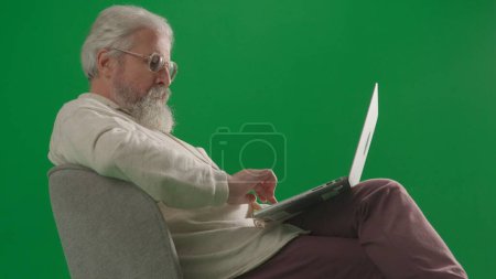 Photo for Pensioner everyday life creative advertisement concept. Portrait of aged bearded man on chroma key green screen background. Full shot of senior man sitting on a chair working on laptop. - Royalty Free Image