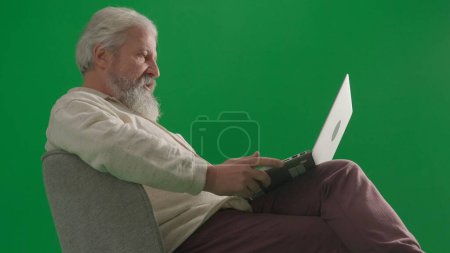 Téléchargez les photos : Pensionné vie quotidienne concept publicitaire créatif. Portrait d'un homme barbu âgé sur fond vert chromé. Plan complet de l'homme âgé assis sur une chaise travaillant sur un ordinateur portable. - en image libre de droit