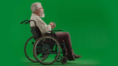 Photo for Pensioner everyday life creative advertisement concept. Portrait of disabled man isolated on chroma key green screen full shot. Senior bearded man sitting in wheelchair holding mug and looking away. - Royalty Free Image