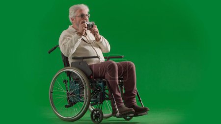 Photo for Pensioner everyday life creative advertisement concept. Portrait of disabled man isolated on chroma key green screen full shot. Senior bearded man sitting in wheelchair holding old film camera. - Royalty Free Image