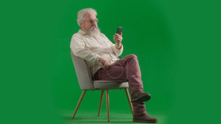 Téléchargez les photos : Pensionné vie quotidienne concept publicitaire créatif. Portrait d'un homme barbu âgé sur fond vert chromé. Plan complet de l'homme âgé assis sur une chaise regardant la vidéo sur smartphone. - en image libre de droit