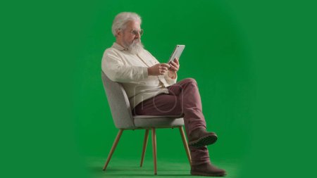 Téléchargez les photos : Pensionné vie quotidienne concept publicitaire créatif. Portrait d'un homme barbu âgé sur fond vert chromé. Plan complet de l'homme âgé assis sur une chaise livre de lecture sur tablette. - en image libre de droit