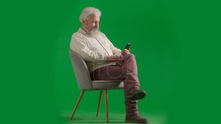 Téléchargez les photos : Pensionné vie quotidienne concept publicitaire créatif. Portrait d'un homme barbu âgé sur fond vert chromé. Plan complet de l'homme âgé assis sur une chaise tenant smartphone, lire les nouvelles. - en image libre de droit