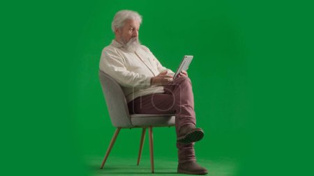 Téléchargez les photos : Pensionné vie quotidienne concept publicitaire créatif. Portrait d'un homme barbu âgé sur fond vert chromé. Plan complet de l'homme âgé assis sur une chaise tenant tablette regarder les médias. - en image libre de droit
