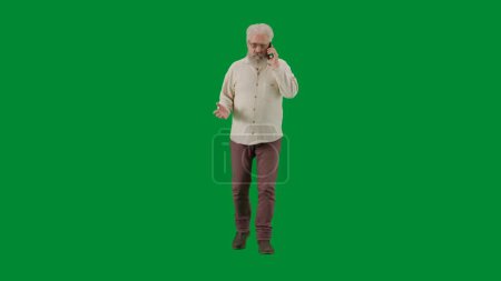 Téléchargez les photos : Pensionné vie quotidienne concept publicitaire créatif. Portrait d'un homme barbu âgé sur fond vert chromé. Plan complet homme âgé marchant et parlant sur smartphone, vue de face. - en image libre de droit