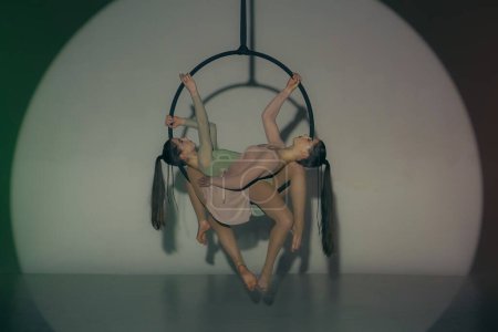Téléchargez les photos : Chorégraphie moderne et acrobaties concept publicitaire créatif. Deux gymnastes isolées sur fond de studio vert. Filles danseurs aériens montrant élément en miroir sur anneau avec des sangles. - en image libre de droit