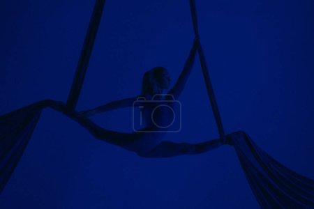 Téléchargez les photos : Chorégraphie moderne et acrobaties concept publicitaire créatif. Silhouette d'artiste féminine isolée sur fond bleu néon studio. Fille danseuse aérienne effectuant élément split volant sur soie aérienne. - en image libre de droit