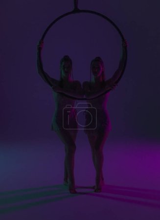 Foto de Coreografía moderna y acrobacia concepto de publicidad creativa. Siluetas de dos gimnastas sobre fondo púrpura estudio de neón. Chicas bailarinas aéreas posando junto al anillo de aire - Imagen libre de derechos