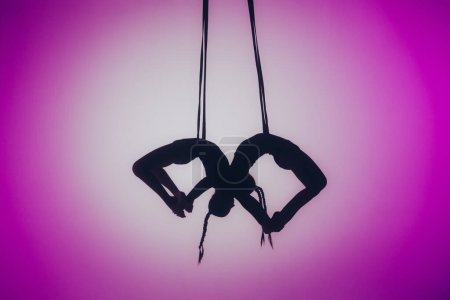 Téléchargez les photos : Chorégraphie moderne et acrobatie concept créatif. Silhouette de deux acrobates femelles sur fond néon rose avec un projecteur au centre. Les acrobates femelles exécutent des éléments acrobatiques sur les cordes - en image libre de droit
