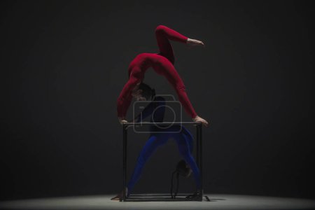 Téléchargez les photos : Chorégraphie moderne et acrobaties concept publicitaire créatif. Portrait de deux acrobates femelles isolées sur fond noir. Filles danseuses aériennes en costume bleu rouge montrant l'élément sur un cube. - en image libre de droit
