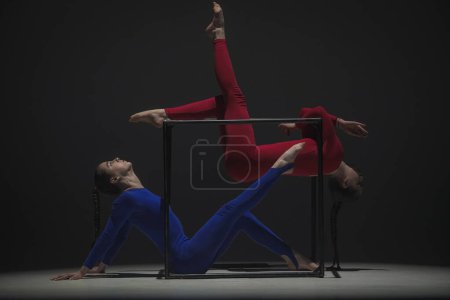 Téléchargez les photos : Chorégraphie moderne et acrobaties concept publicitaire créatif. Portrait de deux acrobates femelles isolées sur fond noir. Filles danseuses aériennes en costume bleu rouge montrant l'élément sur un cube. - en image libre de droit