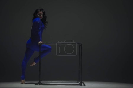 Téléchargez les photos : Chorégraphie moderne et acrobaties concept publicitaire créatif. Portrait d'acrobate femelle isolé sur fond noir. Femme danseuse de gymnastique en body étincelant bleu montrant l'élément sur un cube. - en image libre de droit