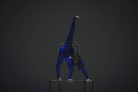 Téléchargez les photos : Chorégraphie moderne et acrobaties concept publicitaire créatif. Portrait d'acrobate femelle isolé sur fond noir. Femme danseuse de gymnastique en body étincelant bleu montrant l'élément sur un cube. - en image libre de droit