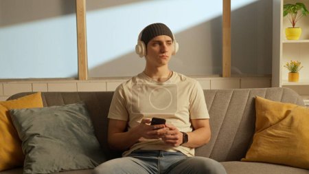 Photo for Music and human emotions creative advertisement concept. Portrait of young person in the room sitting on the couch. Man in headphones listening to music on smartphone. - Royalty Free Image