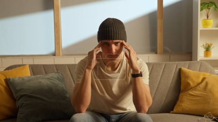 Photo for Human emotion creative concept advertising. Portrait of a distressed young man in a room sitting on a couch. Man in a hat sitting on a sofa and holding his hands on his head - Royalty Free Image