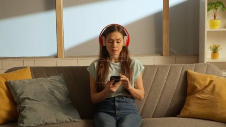 Musik und menschliche Emotionen kreatives Werbekonzept. Porträt eines jungen Menschen im Zimmer, der auf der Couch sitzt. Frau mit Kopfhörer hört Musik auf Smartphone.
