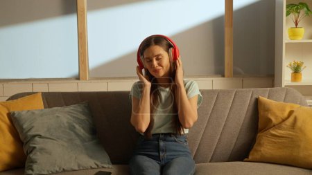 Photo for Music and human emotion creative advertising concept. Woman sitting on a couch listening to music with big red headphones - Royalty Free Image