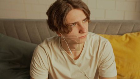 Musik und menschliche Emotionen kreatives Werbekonzept. Porträt eines jungen Menschen im Zimmer, der auf der Couch sitzt. Mann mit Kopfhörer hört Musik auf Smartphone.
