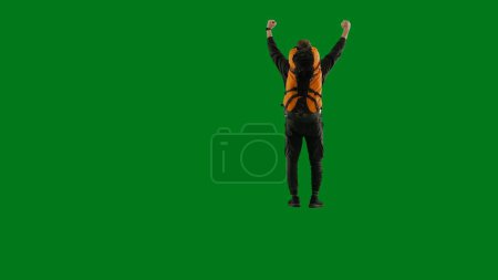 Photo for A young tourist joyfully makes a victory gesture, raising his hands in the air. A man with a large backpack on his back stands full-length in the studio on a green screen. Back view - Royalty Free Image