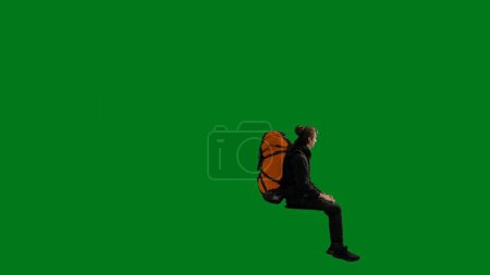Photo for Side view of a male traveler with a backpack on his back, sitting on an elevated position in a studio on a green screen. The man sits with his legs overhanging, puts his hand to his eyes and stares - Royalty Free Image