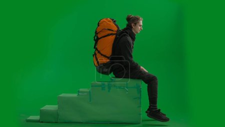 Photo for Side view of a male traveler with a backpack on his back, sitting on an elevated position in a studio on a green screen. The man sits with his legs overhanging, puts his hand to his eyes and stares - Royalty Free Image