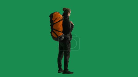 Téléchargez les photos : Vue demi-tournée d'un voyageur masculin avec un grand sac à dos de randonnée sur le dos. L'homme est debout sur toute la longueur et profite de la vue. Randonneur en studio sur écran vert - en image libre de droit