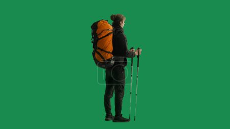 Photo for Half-turned view of a male traveler with a large hiking backpack on his back and trekking poles in his hands. The man is standing full-length and enjoying the view. Hiker in studio on green screen - Royalty Free Image