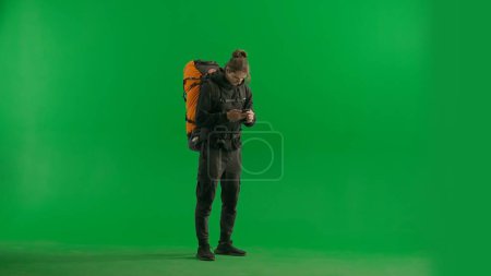 Photo for Male traveler texting on his smartphone. A man with a hiking backpack and with a smartphone in the studio on the green screen. searching for information, communicating on the Internet - Royalty Free Image