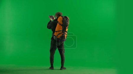 Téléchargez les photos : Un voyageur prend des photos avec un appareil photo en randonnée. Un touriste avec un sac à dos sur le dos se tient toute la longueur dans un studio sur un écran vert. Concept de voyage, repos actif, randonnée - en image libre de droit
