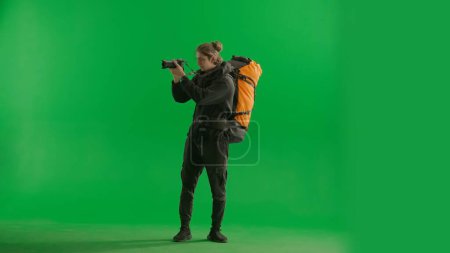 Photo for A male traveler takes pictures with a camera while hiking. A tourist with a backpack on his back stands full length in a studio on a green screen. Concept of travel, active rest, hiking - Royalty Free Image