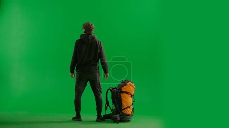Téléchargez les photos : Vue arrière d'un voyageur masculin avec un grand sac à dos de randonnée debout à côté de lui. Homme debout à pleine hauteur et profitant de la vue. Randonneur en studio sur écran vert - en image libre de droit
