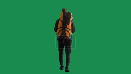 Téléchargez les photos : Un touriste masculin avec un sac à dos sur le dos se promène dans le studio sur un écran vert. Randonnée trek repos voyage concept. Vue arrière - en image libre de droit