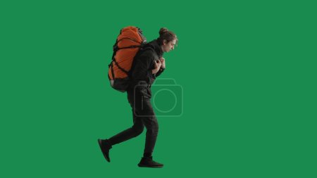 Photo for Side view of a tired hiker in full length walking with a heavy backpack on a green screen. A man wipes sweat from his forehead with his hand. Concept of outdoor adventure, tiredness and fatigue - Royalty Free Image