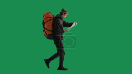 Téléchargez les photos : Un touriste masculin étudie une carte de la région en randonnée. Un voyageur homme en pleine longueur avec un sac à dos sur le dos marchant dans le studio sur l'écran vert - en image libre de droit