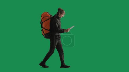 Foto de Un turista masculino estudia un mapa de la zona mientras hace senderismo. Un hombre viajero en longitud completa con una mochila en su espalda caminando en el estudio en la pantalla verde - Imagen libre de derechos