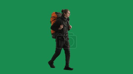Téléchargez les photos : Un touriste masculin avec un sac à dos sur le dos se promène dans le studio sur un écran vert. Randonnée trek repos voyage concept. Vue latérale - en image libre de droit