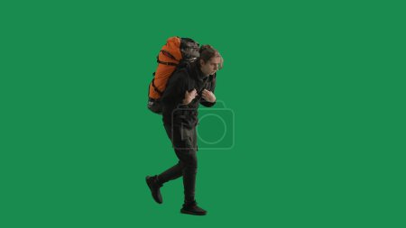 Téléchargez les photos : Vue latérale d'un randonneur fatigué en pleine longueur marchant avec un lourd sac à dos sur un écran vert. Un homme essuie la sueur de son front avec sa main. Concept d'aventure en plein air, fatigue et fatigue - en image libre de droit