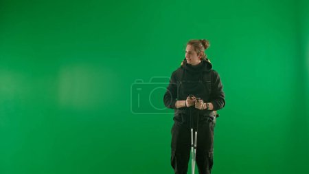 Photo for Male traveler in black tracksuit with trekking poles in hands on green background in studio. The man is standing and looking away - Royalty Free Image