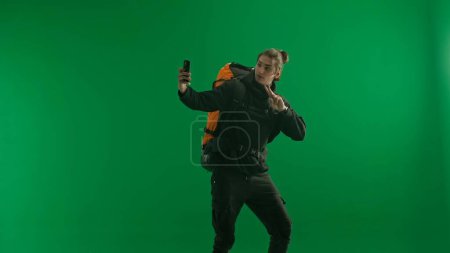 Photo for A male traveler with a backpack on his back on a green screen. The man takes selfies and shows a victory gesture with two fingers - Royalty Free Image