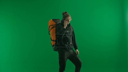Foto de Un viajero masculino con una mochila en la espalda en una pantalla verde. El hombre está hablando en su teléfono inteligente, comunicándose, compartiendo impresiones. - Imagen libre de derechos