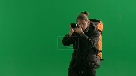 Photo for A male traveler takes pictures with a camera while hiking. A tourist with a backpack on his back stands in a studio on a green screen. Concept of travel, active rest, hiking - Royalty Free Image