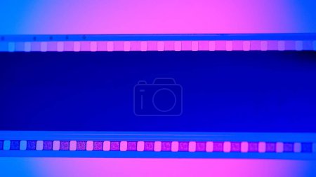 Téléchargez les photos : Bande vierge non développée de film sur fond bleu, éclairée par la lumière rose néon en gros plan. Cadre de diapositive de film 35mm. Cinéma ou cadres photo. Long, cadre de bande de film rétro. Espace de copie - en image libre de droit