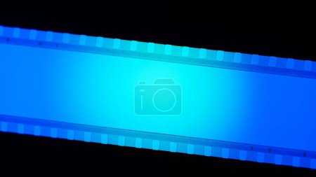 Téléchargez les photos : Bande bleue de film, éclairée de lumière circulaire sur fond noir de près. Bande de cinéma sur fond noir. Cadre de diapositive de film 35mm. Cinéma ou cadres photo. Long, cadre de bande de film rétro - en image libre de droit