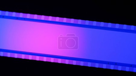Foto de Banda de película azul, iluminada con luz circular rosa sobre fondo negro de cerca. Cinema filmstrip sobre fondo negro. Marco de diapositiva de película de 35 mm. Cinema o marcos de fotos. Marco de tira de película largo y retro - Imagen libre de derechos